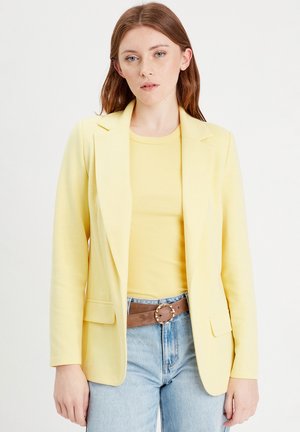 Gul blazer i blødt stof med skræddersyet pasform, hakkrave og to forlommer, kombineret med en gul top og blå jeans.