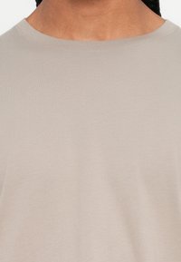 Close-up van een persoon die een lichtbeige crewneckshirt draagt, met zicht op de nek en bovenborst.