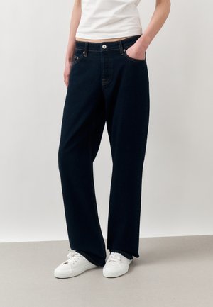 Person som har på seg løse mørkeblå jeans, hvite joggesko og en hvit topp, står med en hånd i lommen mot en ensfarget bakgrunn.