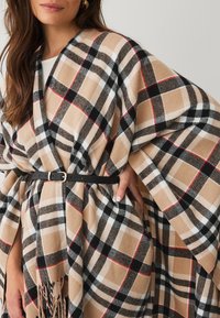 Beige and black plaid shawl with red accents, featuring a fringed hem and secured with a black belt. Soft, textured fabric drapes elegantly.