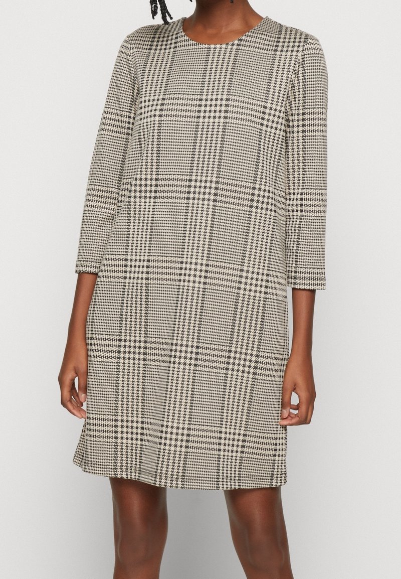 Femme portant une robe beige et noire à motif pied-de-poule, longueur genou, manches 3/4 et encolure ronde.