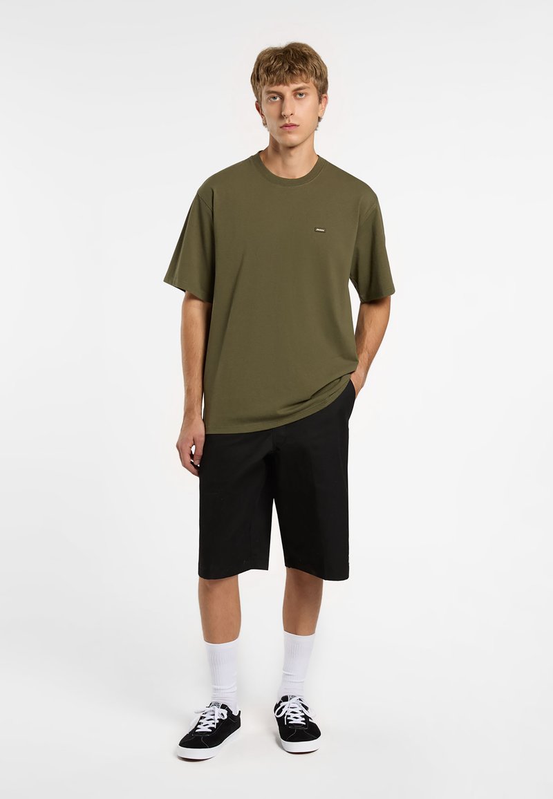 T-shirt verde oliva con colletto rotondo e maniche corte, abbinato a pantaloni corti neri fino al ginocchio. Il modello indossa calze bianche e sneakers nere.