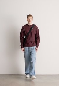 Bordó pulóver, laza szabású világoskék farmernadrág, fehér tornacipő és fekete keresztpántos táska. Modell semleges háttér előtt sétál.