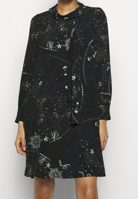 Černé šaty s kosmickým potiskem, který zobrazuje hvězdy a nebeské motivy. Měkká tkanina, dlouhé rukávy a detail vázání u krku.