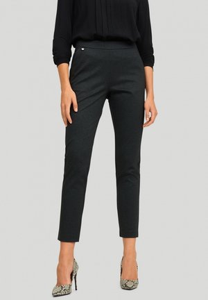 Donna che indossa pantaloni neri aderenti alla caviglia con un leggero motivo a quadri, abbinati a una blusa nera a maniche lunghe e tacchi alti in stampa pitone.