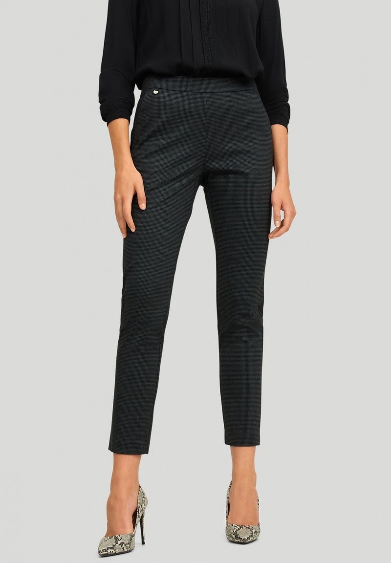 Femme portant un pantalon noir ajusté jusqu'aux chevilles avec un motif à carreaux subtil, associé à un chemisier noir à manches longues et des talons hauts en cuir de serpent.