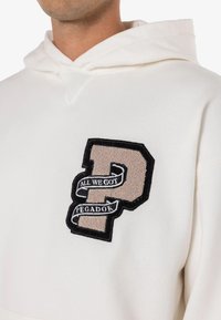 Camisola branca com capuz ajustável, apresentando uma grande letra "P" bordada em bege e preto, realçada com uma fita que diz "ALL WE GOT PEGADOR."