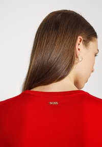 Maglione rosso a maglia con scollo rotondo e un piccolo tag con logo dorato sul retro. Texture liscia e design aderente.