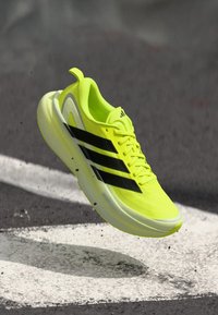 Helles gelbes Sportschuh aus synthetischem Material, mit drei schwarzen Streifen, einem schlanken Design und einer leichten Sohle.
