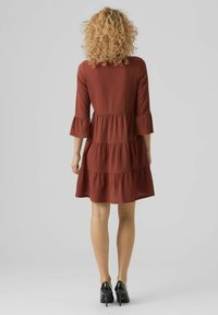 Vestido de color óxido con mangas tres cuartos, diseño en capas, tela texturizada y puños con volantes. Llevado con zapatos de tacón alto negros.