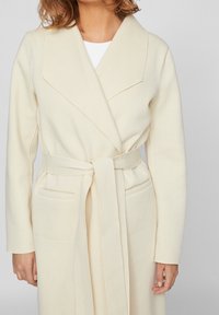 Bata cruzada color crema con un amplio cuello, mangas largas, cinturón anudado y dos bolsillos delanteros. Fabricada con una tela suave y texturizada.