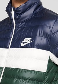 Nike pufferjacka med hög krage, med horisontella ränder i marinblått, vitt och grönt. Logotyp på vänster bröst med dragkedjestängning.
