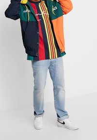 Flerfärgad vindjacka med marinblå, grön och orange paneler; vit grafik på bröstet. Randig skjorta under; ljusblå jeans; vita sneakers.