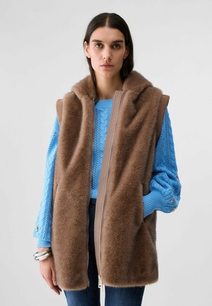 Donna con un gilet senza maniche marrone in pelliccia sopra un maglione di lana blu e jeans scuri, in piedi davanti a uno sfondo chiaro e uniforme.