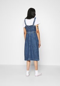 Niebieska dżinsowa sukienka ogrodniczka z regulowanymi ramiączkami, dopasowanym gorsetem i spódnicą sięgającą do kolan. Noszona na białą koszulkę i zestawiona z białymi trampkami.