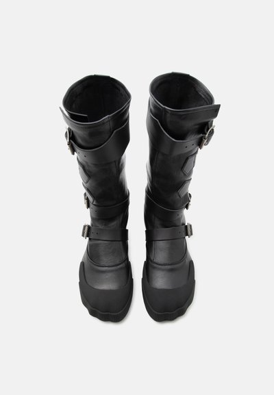 Ψηλές μαύρες δερμάτινες μπότες με πολλούς ρυθμιζόμενους ιμάντες και ενισχυμένες άκρες στο μπροστινό μέρος, παρουσιασμένες όρθιες σε λευκό φόντο.