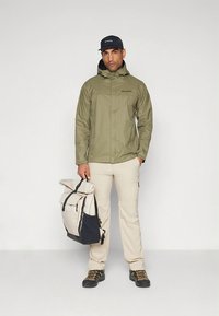 Giacca impermeabile verde oliva, pantaloni beige, scarponi da trekking neri, con uno zaino beige e blu navy, e un berretto nero.