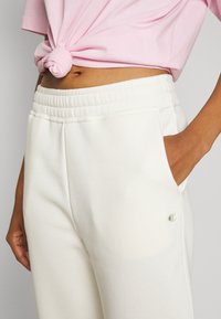 Hvite joggebukser laget av mykt stoff, med elastisk midjebånd, sidelommer og en liten logo på venstre bein.