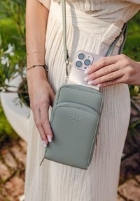 Light green, textured leather crossbody bag with dual zippered compartments, silver logo, and a strap. A smartphone is partially inserted.