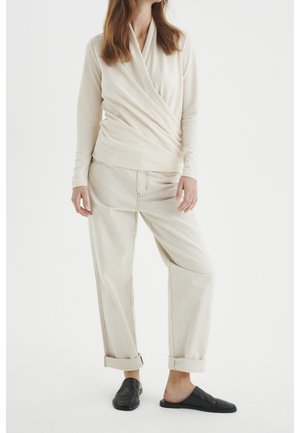 Vrouw draagt een crèmekleurige lange-mouw wikkel top, lichtbeige opgerolde broek en zwarte slip-on loafers, staand tegen een witte achtergrond.