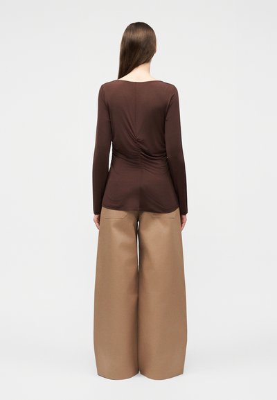 Γυναίκα με μακριά καστανά μαλλιά, ντυμένη με στενό καφέ μακρυμάνικο τοπ και φαρδιά μπεζ παντελόνια, φαίνεται από πίσω σε απλό φόντο.