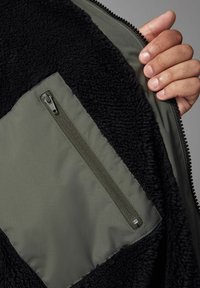 Schwarze Fleecejacke mit gefüttertem Innenbereich, der einen olivgrünen Stoff mit einer Reißverschlusstasche und strukturiertem schwarzen Fleece für Wärme und Komfort zeigt.