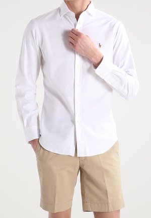 Mann trägt ein weißes Hemd mit Knöpfen und kleinem besticktem Logo sowie beige Shorts und richtet den Hemdkragen mit einer Hand.