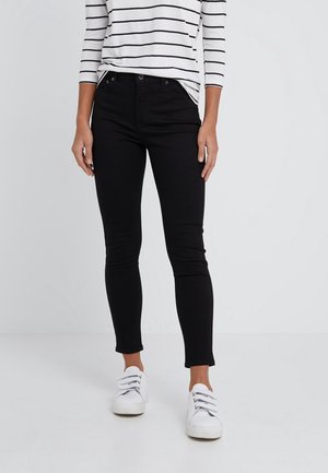 Kvinde iført sorte slim-fit bukser, hvide sneakers og en hvid langærmet top med sorte vandrette striber, stående på en ensfarvet baggrund.