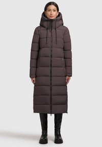 Long, padded coat in dark brown with a quilted design, zip closure, adjustable hood, and side zippers. Paired with black knee-high boots.