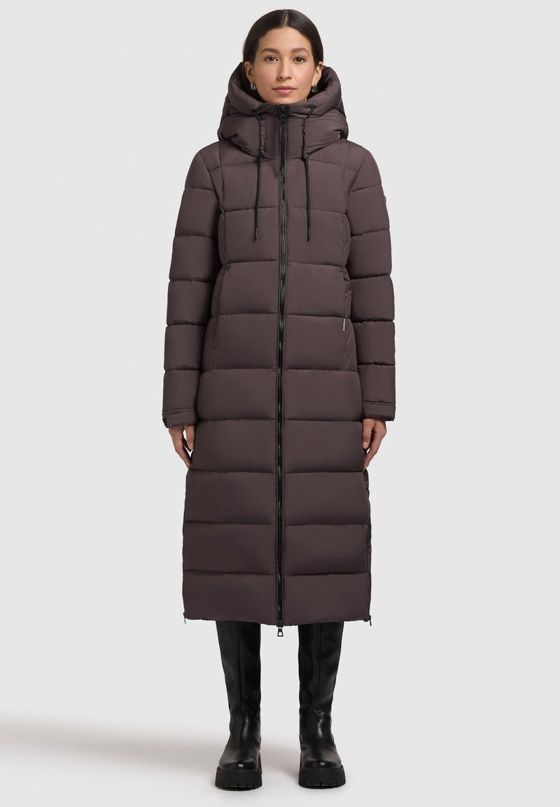 Long, padded coat in dark brown with a quilted design, zip closure, adjustable hood, and side zippers. Paired with black knee-high boots.