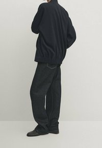 Henkilö, jolla on yllä löysä musta takki, tummat väljät farkut ja mustat loafersit, seisoo yksinkertaista vaaleaa taustaa vasten.