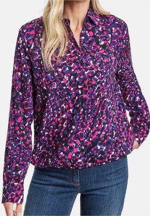Kobieta w koszuli z długim rękawem zapinanej na guziki z abstrakcyjnym wzorem kropek w kolorach różowym, fioletowym i czarnym, w połączeniu z niebieskimi dżinsami.