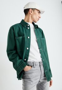 Πράσινο τζιν μπουφάν με κουμπιά σnap και τσέπες, φορεμένο πάνω από μια λευκή μπλούζα και γκρι παντελόνι, σε απλό φόντο. Συμπεριλαμβάνεται καπέλο.