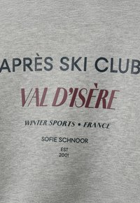 Grijze sweatshirt met zwarte en maroon tekst: "APRÈS SKI CLUB VAL D’ISÈRE WINTERSPORTEN • FRANKRIJK" en "SOFIE SCHNOOR EST 2001."