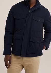 Man i en marinblå zip-up jacka med fyra framfickor och beige byxor, hand i fickan, mot en plain beige bakgrund.