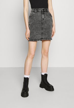 Femme portant une jupe en jean noire délavée, un débardeur noir, des chaussettes blanches et des bottines à plateforme noires à lacets, debout sur un fond uni.