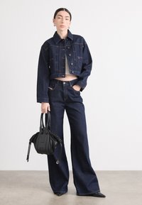 Mørk denimjakke med en cropped design og knaplukning, parret med vide, mørke jeans. Sort håndtaske med strukturerte detaljer.