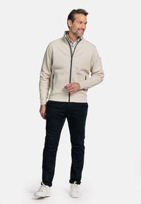 Beige zip-up jack met een staande kraag, zwarte ritssluiting en zijzakken, gecombineerd met donkere broek en witte sneakers. Eenvoudig ontwerp, gladde textuur.