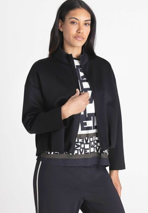 Woman wearing a black zip-up jacket over a white graphic shirt and black pants with a side stripe, looking at the camera with hand near the zipper.