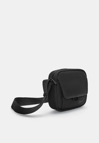 Sort stof crossbody tas med rektangulær form, lynlåslukning, justerbar strop og en frontklap med logo-detalje.