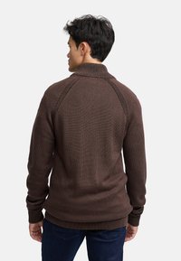 Brązowy sweter dzianinowy z wysokim kołnierzem, prążkowanymi mankietami i wyraźnymi szwami na ramionach. Gładka faktura uzupełnia dopasowany krój.