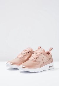 Ett par roséguld Nike-sneakers med vita sulor, övre del i nät och läder, samt snörning, visade mot en enkel vit bakgrund.
