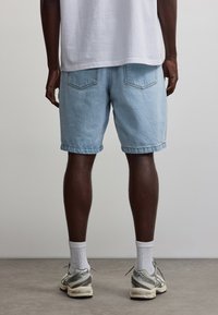 Person steht drinnen mit dem Rücken zur Kamera, trägt ein weißes Shirt, hellblaue Jeansshorts, weiße Crew-Socken und graue Sneaker auf grauem Boden.