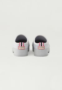Biele slip-on topánky s masívnou šedou podrážkou a červeným a čiernym prúžkom na päte. Obsahujú označení značky v červenej farbe na zadnej strane.