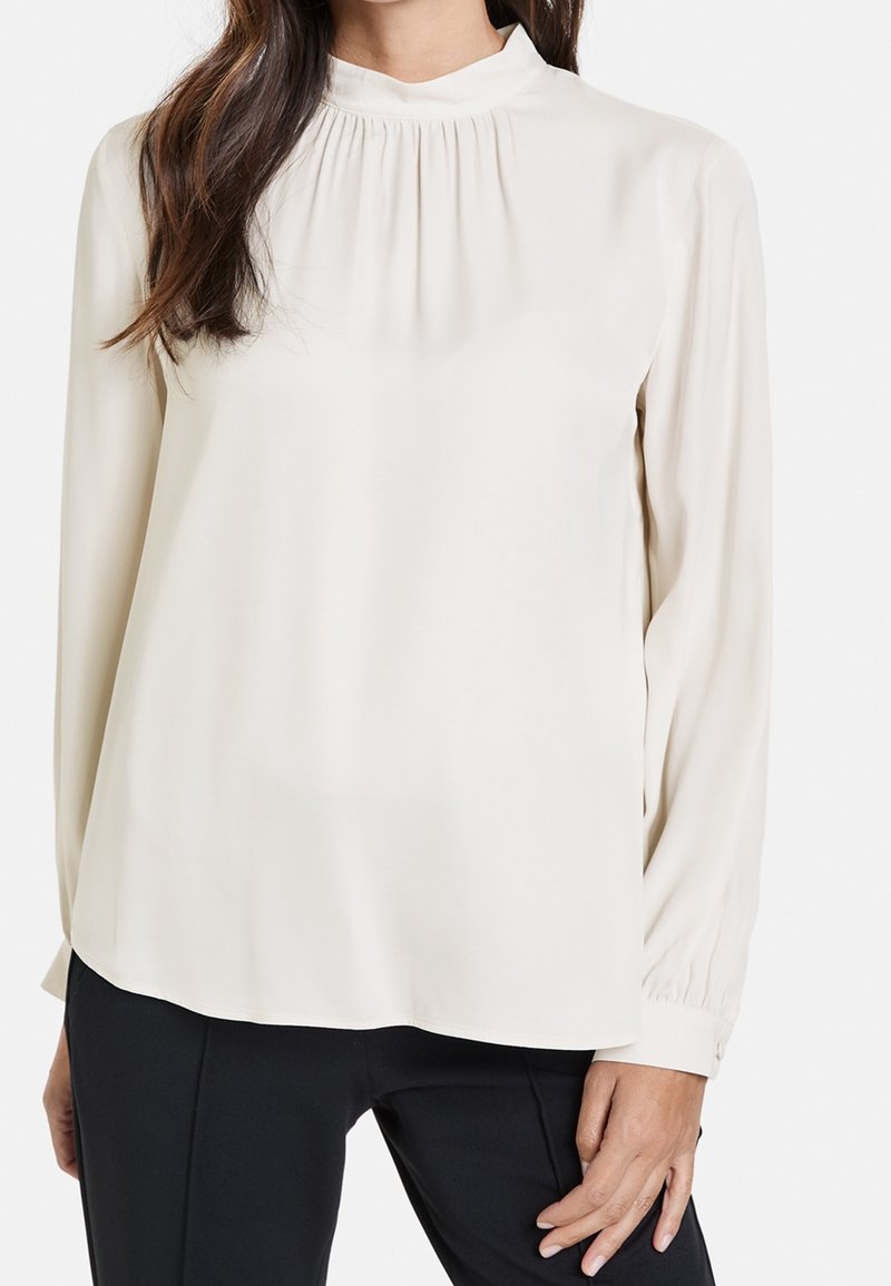 Blusa leggera color crema con un collo alto, dettagli arricciati al giogo e maniche lunghe. Tessuto liscio, taglio dritto, abbinata a pantaloni scuri.