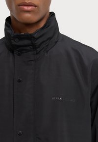 Černá bunda s vysokým límcem, s lesklým povrchem a zapínáním na patentky. Logo "Armani Exchange" diskrétně umístěné na přední straně.