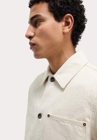Light beige jacket with a textured fabric, featuring a collared neckline, black buttons, and a chest pocket with matching accents.