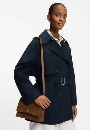 Cappotto blu navy con collo ampio, bottoni doppi e cintura. Borsa in suede color cuoio con dettaglio a chiusura, indossata sulla spalla.