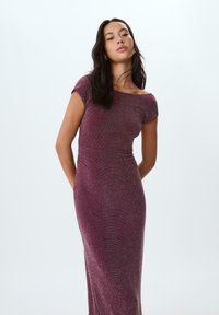 Donna con lunghi capelli scuri che indossa un elegante abito viola aderente con spalline scoperte, in piedi con le mani dietro la schiena su uno sfondo semplice.