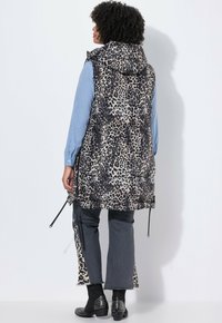 Wattierte Weste mit Leopardenmuster, Kapuze und Kordelzugsaum. Kombiniert mit einem blau gestreiften Hemd und dunklen Jeans, die tierische Druckakzente aufweisen.